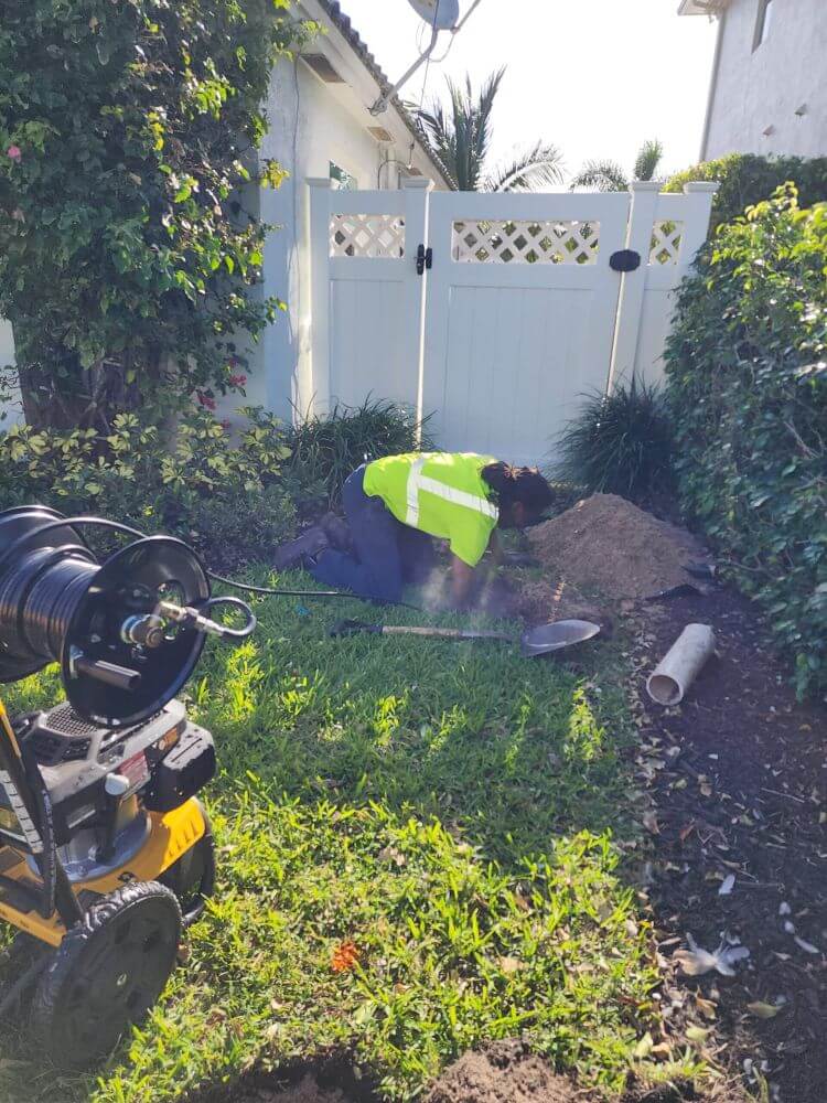 Tip Top Plumbing technician performs hydro jetting at a Pembroke Pines residence using a high-pressure machine to clear underground drain lines. The equipment hose is guided into an exposed sewer access point in the homeowner’s yard.