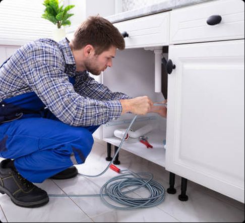 Technician in Plantation FL performing kitchen drain cleaning under a white cabinet with a plumbing snake, showcasing Tip Top Plumbing & Restoration's professional services.